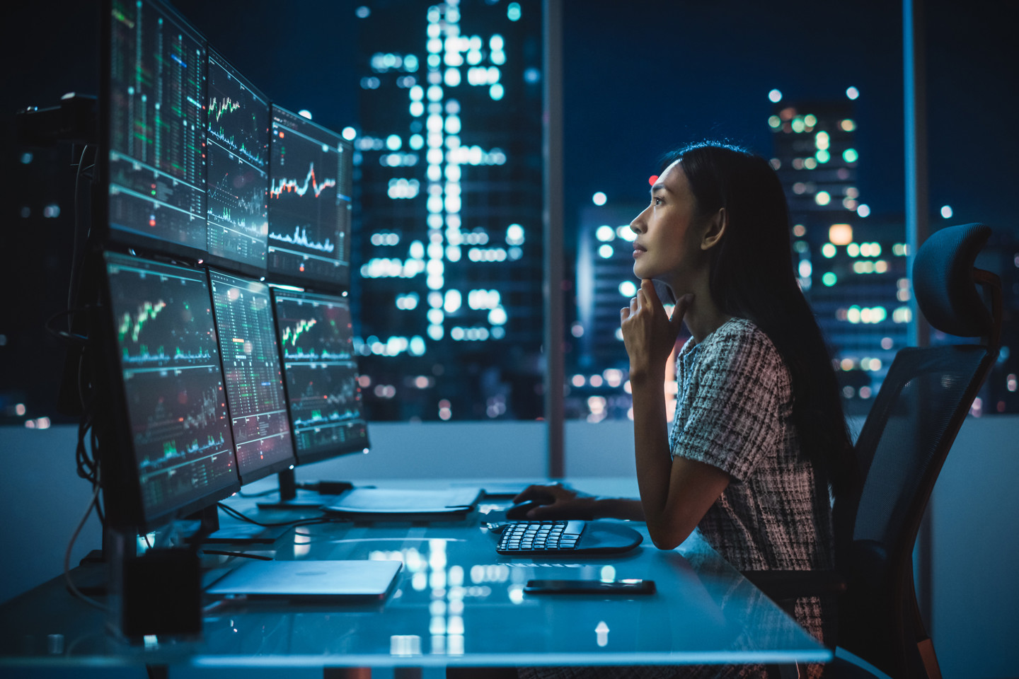 Woman sat at her office desk looking at monitors with financial data - city buildings can be seen in the background